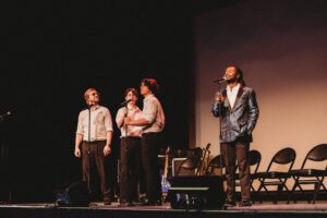 A group of men singing on stage with one man holding a microphone.